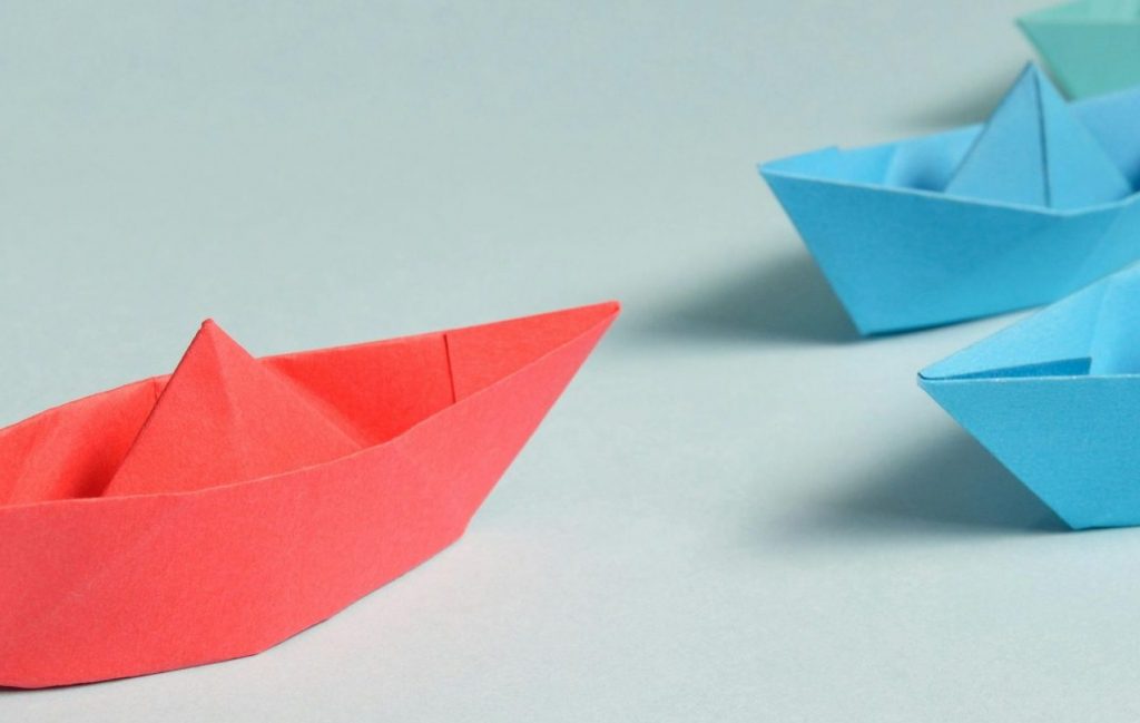 Ein großes rotes Papierschiff führt viele kleine blaue Papierschiffe an – Symbol für erfolgreiche Führung.